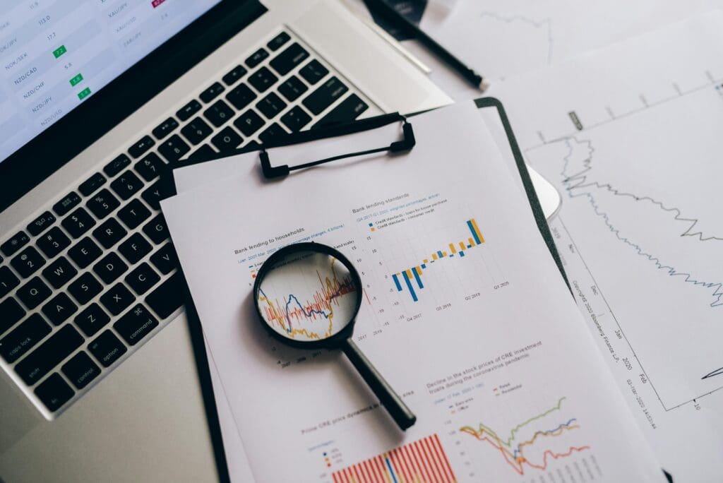 Data analysis workspace with laptop and financial charts. Magnifying glass focusing on colorful bar graph with blue and orange indicators. Statistical reports and trend line graphs visible on white paper, displaying market performance metrics.
