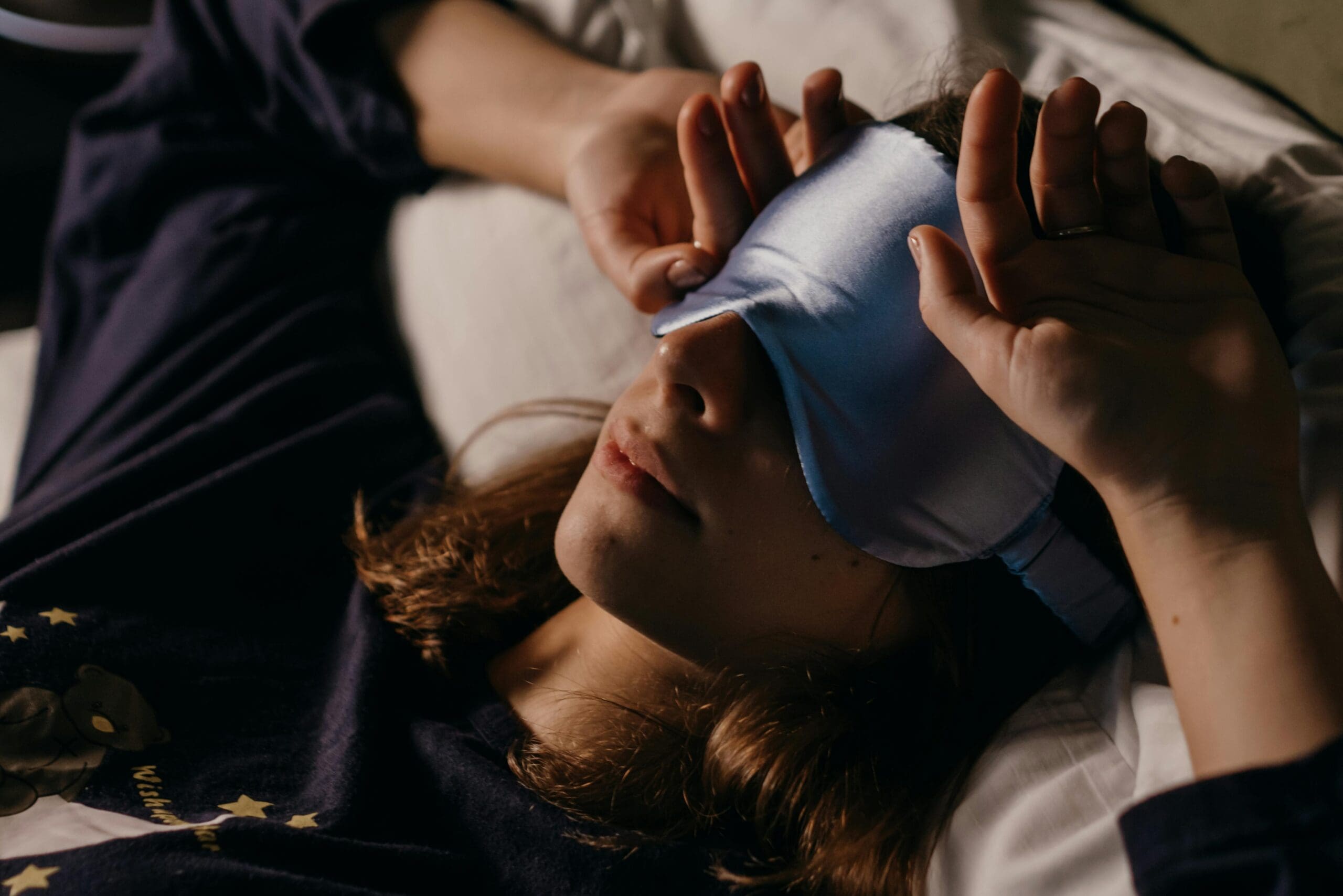Person wearing blue sleep mask lying in bed with white sheets and starry navy blanket. Hands adjusting sleep mask for optimal comfort. Deep sleep optimization for improved rest quality and recovery.