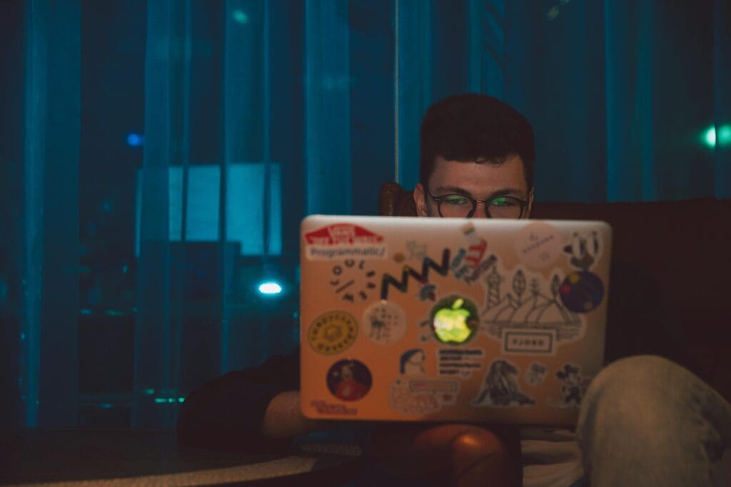 Person with glasses working on MacBook laptop in dimly lit room with blue ambient lighting. Laptop covered with programming and tech stickers including Apple logo glowing in dark. Evening digital work environment showcasing late-night work session and blue light exposure.
