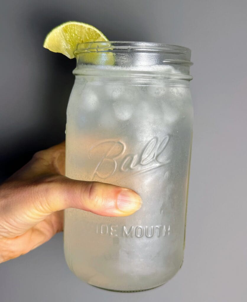 A hand holding a 24-ounce Ball mason jar filled with a clear 'boozeless' margarita made with water, sparkling water, fresh lime juice, salt and ice. A lime wedge garnishes the rim, and condensation appears on the glass, emphasizing hydration.