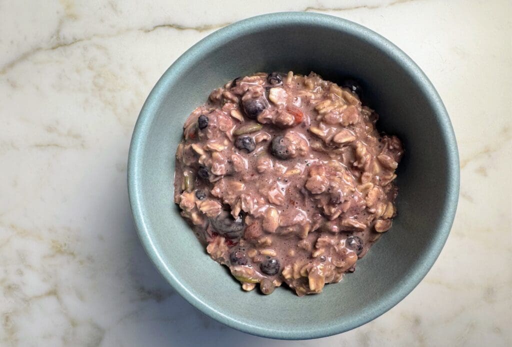 Overhead view of a blue ceramic bowl containing overnight oats mixed with blueberries, showing a creamy purple-brown texture with visible oats and whole blueberries throughout, placed on a white marble surface.