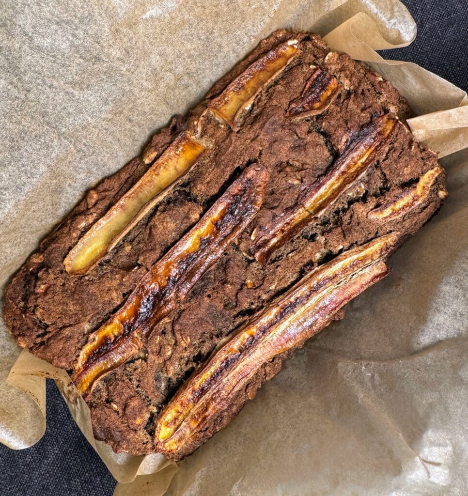 Overhead view of freshly baked vegan banana bread made with buckwheat flour, showing a rich brown color with caramelized banana slices baked into the top surface, sitting on parchment paper.