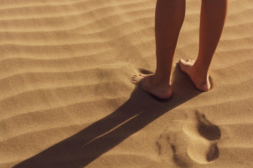 Person walking barefoot on textured sand with visible footprints, demonstrating natural foot movement.
