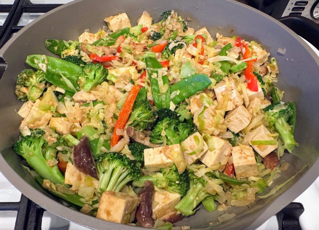 Colorful vegetable stir-fry in large skillet with tofu, broccoli, bell peppers, snap peas, and mushrooms, showing realistic plant-based meal preparation