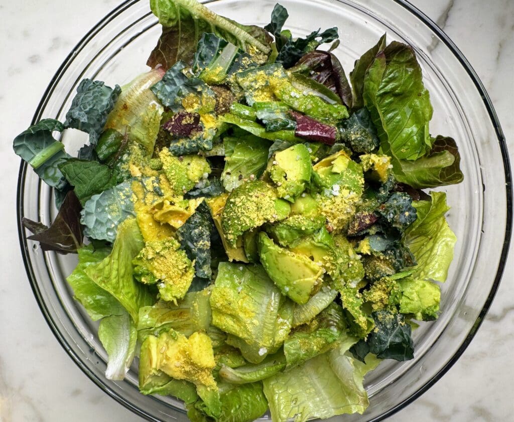 Large glass mixing bowl containing unmixed salad with diverse leafy greens, mashed avocado, and nutritional yeast before tossing