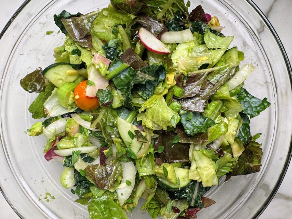 Mixed salad in glass bowl with diverse vegetables, herbs, radishes, cucumber, and cherry tomato coated in avocado and lemon dressing