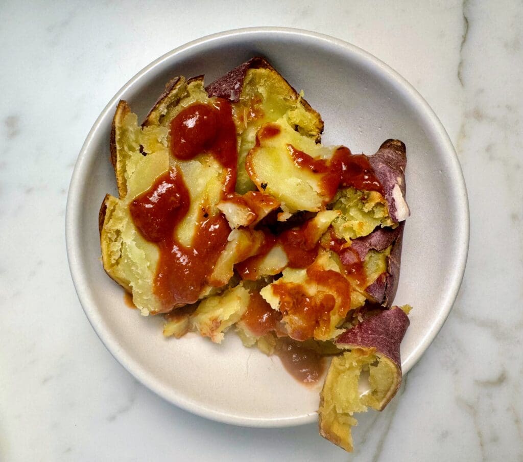 Baked Japanese sweet potato with purple skin and orange flesh topped with ketchup on white plate