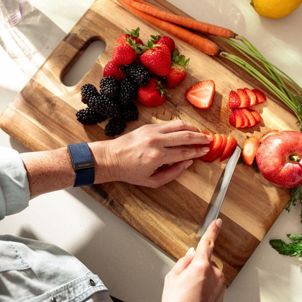 Hands wearing Whoop fitness tracker preparing healthy foods including strawberries, blackberries, and vegetables on wooden cutting board