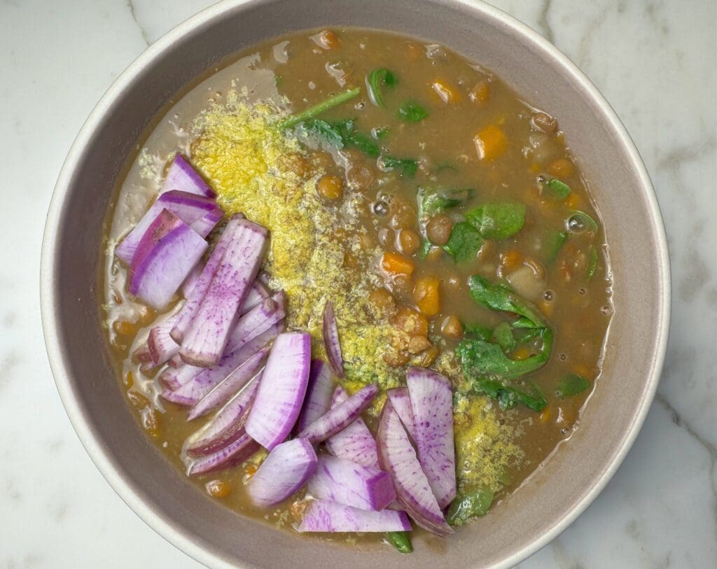 Bowl of lentil soup with sliced radish, nutritional yeast, and spinach demonstrating simple plant-based meal assembly