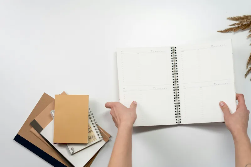 Hands writing in open spiral planner with notebooks on white desk representing daily goal tracking habit