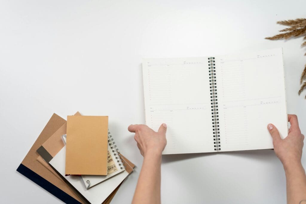 Hands writing in open spiral planner with notebooks on white desk representing daily goal tracking habit