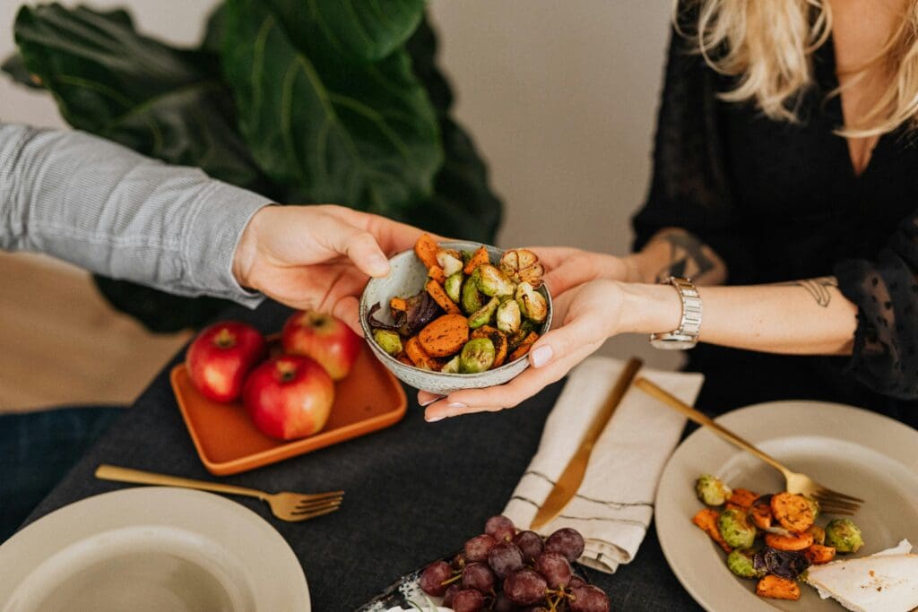 Hands sharing a bowl of colorful roasted vegetables including Brussels sprouts and carrots for healthy Thanksgiving