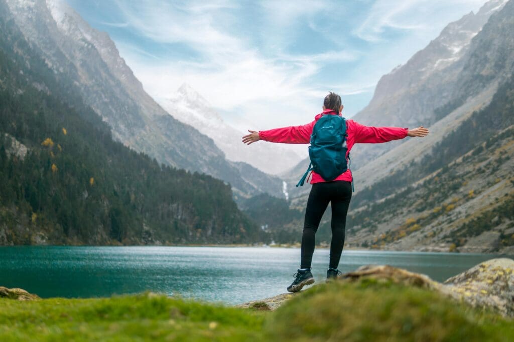 Woman in red jacket standing arms wide open on a rock overlooking a mountain lake, backpack on, facing the mountains.
