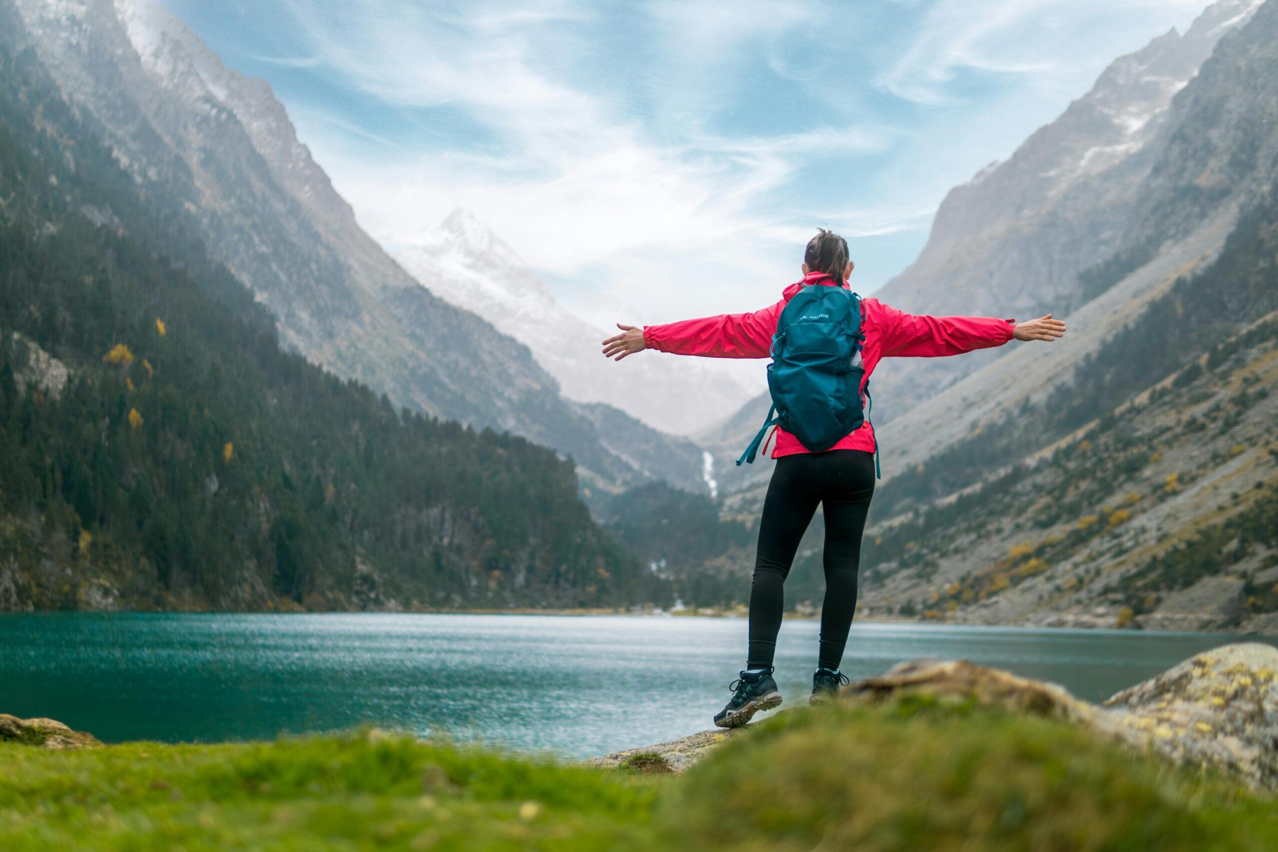 Woman in red jacket standing arms wide open on a rock overlooking a mountain lake, backpack on, facing the mountains.