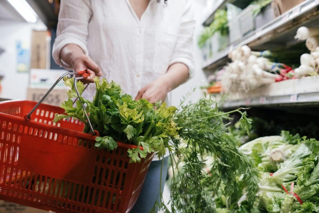 Person holding red grocery basket filled with fresh celery and leafy greens, illustrating whole foods shopping for heart health