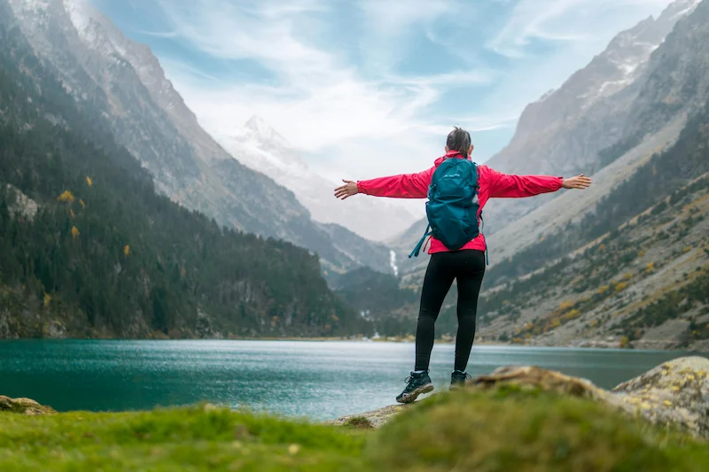 Woman in red jacket standing arms wide open on a rock overlooking a mountain lake, backpack on, facing the mountains.