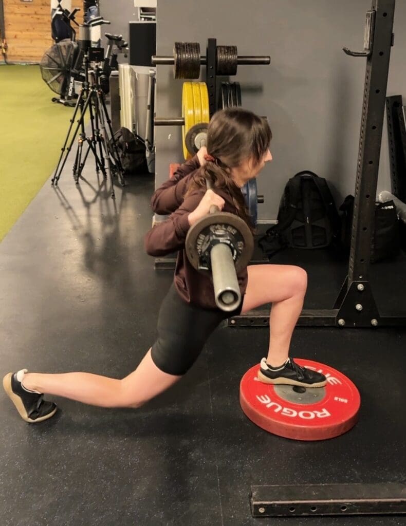 A woman in her 50s performing a barbell deficit reverse lunge with a 45-pound barbell across her shoulders, front foot elevated on a weight plate, in a strength-training gym.