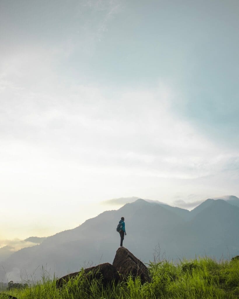 A hiker with a backpack stands on a rock overlooking a misty mountain range at dawn.