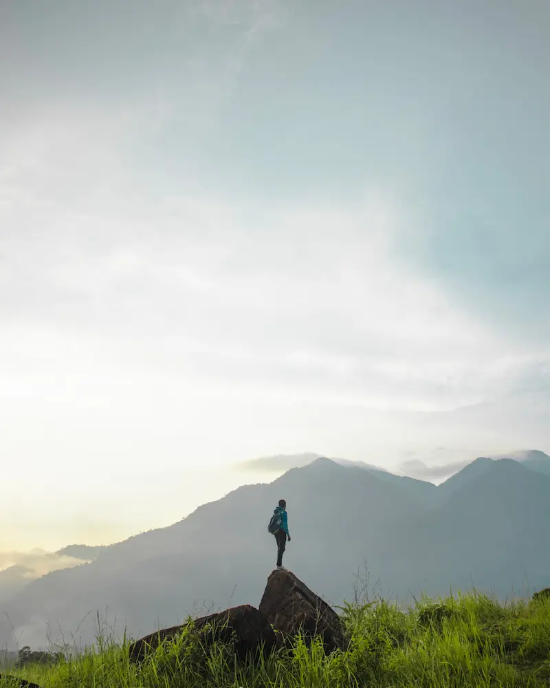 A hiker with a backpack stands on a rock overlooking a misty mountain range at dawn.